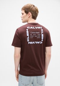 Camiseta de algodón marrón con gran texto blanco en contraste en la parte trasera, que muestra "Calvin Klein" y detalles de dirección en un diseño cuadrado.