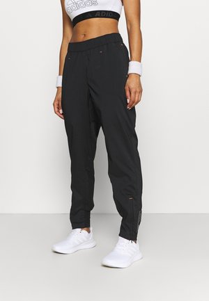 Frau trägt schwarze Adidas-Trainingshose, weißen Sport-BH, weiße Handgelenkbandagen und weiße Sneakers, steht vor einem einfarbigen Hintergrund.