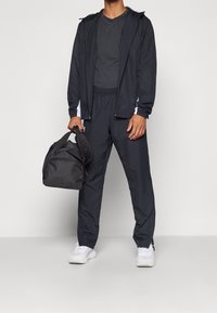 Schwarzes Sporttracksuit mit einer Reißverschlussjacke und Hose mit elastischem Bund, kombiniert mit einer schwarzen Sporttasche und weißen Sneakern.