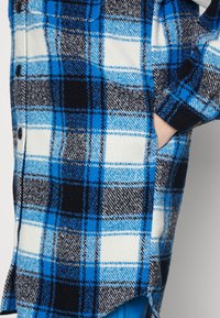 Giacca camicia a quadri blu, neri e bianchi, realizzata in tessuto morbido, con bottoni neri e tasche laterali; presenta un orlo leggermente curvo.