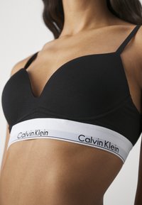 Svarte polstrede bh med tynne stropper, med et hvitt elastisk bånd som viser "Calvin Klein" i svart. Glatt tekstur.