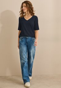 Marineblå V-hals ribbet top og lys blå slidte lige ben jeans, kombineret med hvide sneakers på en neutral baggrund.