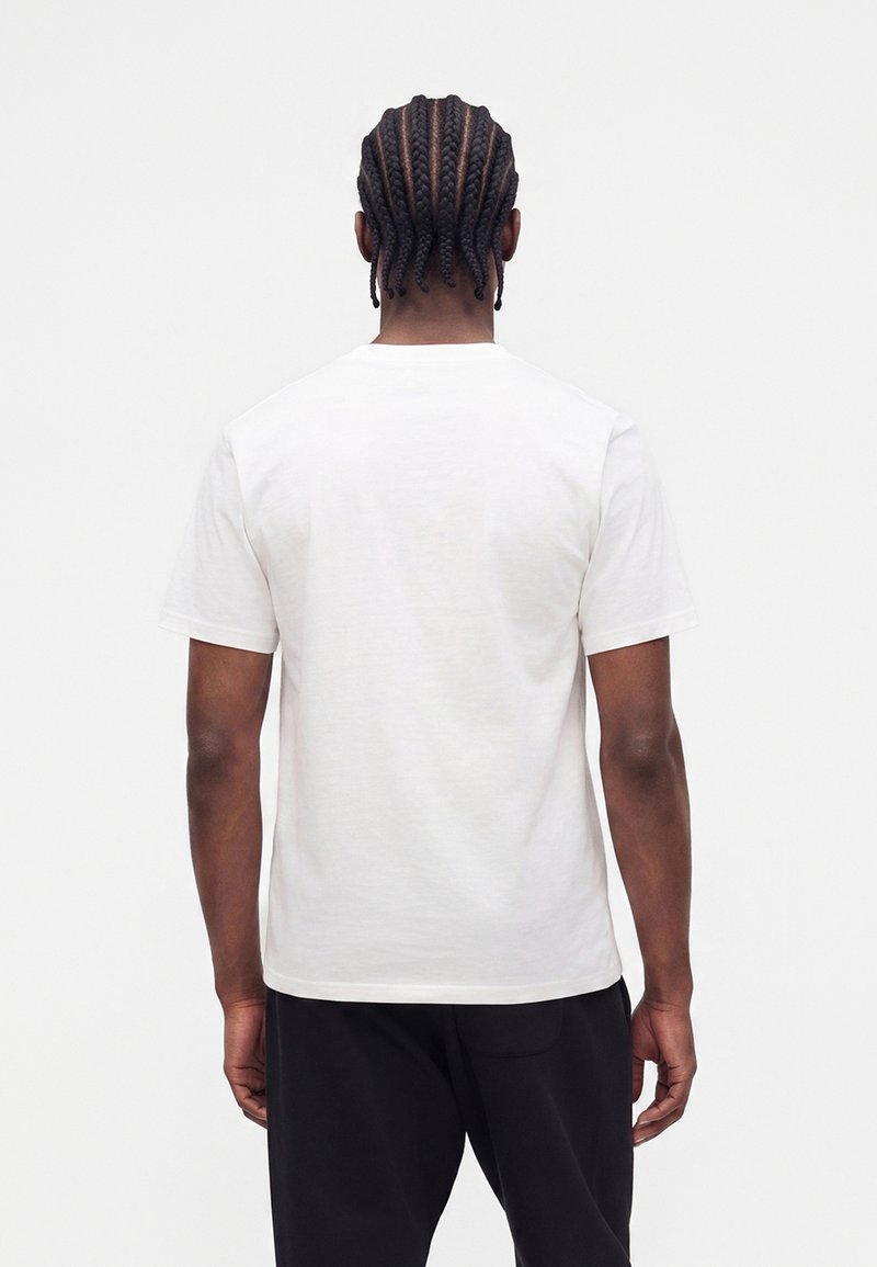 Man with cornrow braids wearing a plain white short-sleeve t-shirt and black pants, standing facing away against a white background.