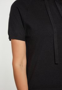 Top nero a maniche corte realizzato in morbido tessuto lavorato a maglia, con orlo arrotondato e dettaglio di coulisse anteriore. Texture liscia con un leggero luccichio.