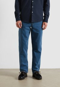 Jeans en denim bleu, coupe droite, lavage moyen, design à cinq poches, accompagnés de chaussures noires avec une semelle texturée et des lacets.