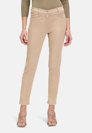 Beige fitted pants with a smooth texture, mid-rise waist, and front pockets, paired with strappy, neutral-toned high heels.
