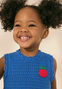 Haut en crochet bleu avec un col en dentelle et un design sans manches. Présente un patch en forme de pomme rouge avec un détail de feuille verte à l'avant.