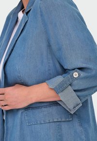 Blazer de mezclilla azul claro con un corte relajado, que presenta mangas enrolladas, cierre de un solo botón y detalle de bolsillo delantero.