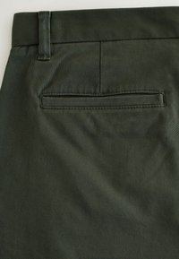 Pantaloni in tessuto verde scuro con tasca posteriore piatta, passanti per cintura, texture liscia e lieve lucentezza. Design su misura con linee pulite.