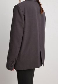 Blazer gris con solapa con muesca, abertura simple en la parte posterior y puños de tres botones. La tela es suave con un aspecto ajustado y estructurado.