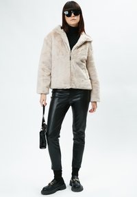 Giacca in pelliccia sintetica beige chiaro con zip e cappuccio, dolcevita nero, pantaloni in pelle sintetica nera lucida e mocassini neri chunky.