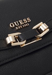 Borsa nera in ecopelle testurizzata con logo in metallo dorato "GUESS EST 1981" e una chiusura decorativa. Design lussuoso e elegante con rifiniture lucide.