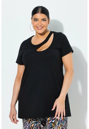 Smilende kvinde med mørkt hår i en glat knold iført sort top med udskæringer, store hoops-øreringe og farverige bukser med zebramønster.