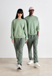 Two models wearing matching light green sweatshirts and patterned green track pants, one with sunglasses and the other with a pink cap.