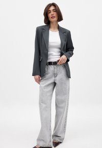 Grå blazer med en glat overflade, parret med lysegrå brede jeans. Hvid top under, sort bælte og mørke sko.