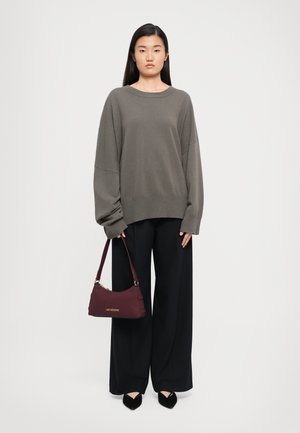 Woman wearing oversized gray sweater and wide-leg black pants, holding small burgundy handbag with gold lettering.