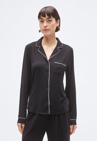 Mustad pikkade varrukatega pidžaamatopp valge äärega, ees nupud ja rinnatasku. Pehme kangas ja lõdvestunud lõige.