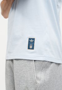 Ανοιχτό μπλε πουκάμισο με το σήμα της Adidas και της Ιταλίας φορεμένο πάνω από γκρι φόρμες με ορατή τσέπη και ραφές.