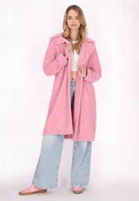 Geacă lungă, pufoasă, de un roz deschis, cu glugă și un design deschis în față. Purtați-o peste un top scurt alb și blugi evazați de culoare albastru deschis.