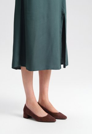 Brązowe zamszowe buty na słupku z ostrym noskiem i niskim wycięciem, w połączeniu z zieloną sukienką z bocznym rozcięciem.