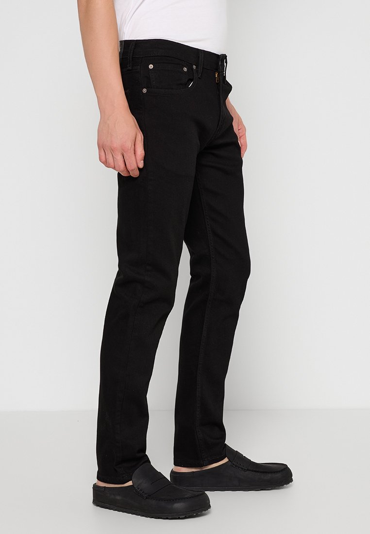 Schwarze Denim-Jeans mit schlanker Passform, fünfw pockets Design, metallenen Nieten und einem Bund mit Gürtelschlaufen, kombiniert mit schwarzen Slippern.
