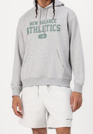 Personne portant un sweat à capuche New Balance Athletics gris clair avec poche avant et un short gris clair assorti avec cordon de serrage et logo.