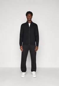 Jaqueta preta com fecho zip na frente e gola alta, combinada com calças cinza escuras. O modelo usa ténis brancos. Textura suave, design elegante.