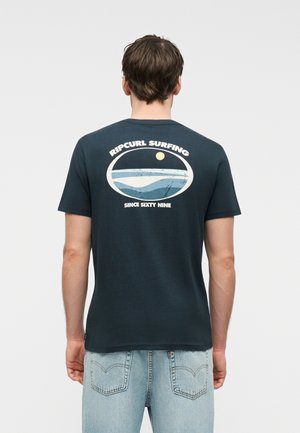 Mees näoga eemal, seljas tumesinine Ripcurl Surfing T-särk, millel ookeani ja päikese kujutis, ning helesinised teksad.
