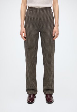 Personne portant un pantalon marron à carreaux à jambes droites et des mocassins en cuir rouge foncé, debout sur un fond blanc uni.