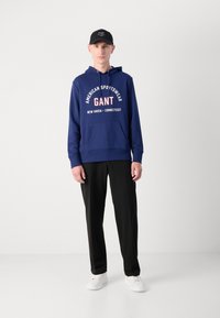 Sudadera azul marino con estampado blanco y rojo, bolsillo canguro; combinada con pantalones negros y zapatillas blancas, finalizada con una gorra negra.