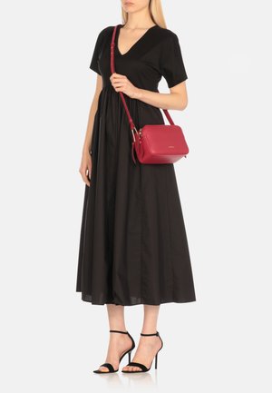 Borsa a tracolla in pelle rossa con forma rettangolare, chiusura con zip e tracolla regolabile. Modella che indossa un abito nero e sandali neri con cinturini.