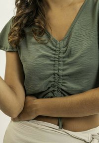 Haut olive vert à manches courtes avec un col en V, présentant des détails froncés à l'avant et un accent noué à la taille. Texture lisse.