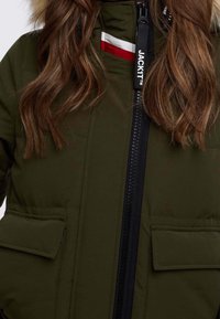 Veste rembourrée vert olive avec fermeture éclair noire, dotée de poches avant et d'une capuche doublée en fourrure. Comprend des bandes accentuées rouges et blanches sur la poitrine.