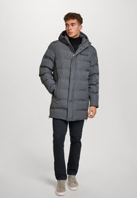 Giubbotto imbottito grigio con cappuccio, chiusura con zip e tasche frontali. Presenta un logo sul petto e polsini a costine, abbinato a pantaloni scuri.