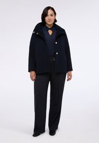 Cappotto in misto lana blu navy con cappuccio e bottoni dorati, abbinato a una blusa blu scuro e pantaloni grigio scuro su misura. Texture liscia, design elegante.