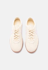 Ζεύγος μπεζ σουέντ και υφασμάτινων αθλητικών παπουτσιών με ροζ λογότυπα και καουτσούκ σόλες, σε θέα από πάνω σε λευκό φόντο.