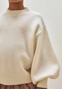 Maglione lavorato a maglia di colore crema, con un collo rotondo alto, orlo a coste e maniche a sbuffo oversize. Texture morbida, design a metà vita e un aspetto accogliente.