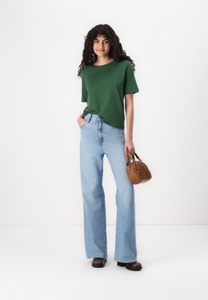 Maglietta verde a maniche corte abbinata a jeans a vita alta di un blu chiaro e a gamba larga. Tiene una piccola borsa marrone con un motivo intrecciato.