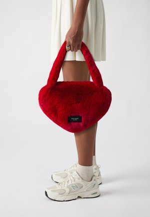 Rød plyss hjertedannet håndtaske med to korte håndtag og en sort label. Model holder tasken, iført en hvid kjole og beige sneakers.