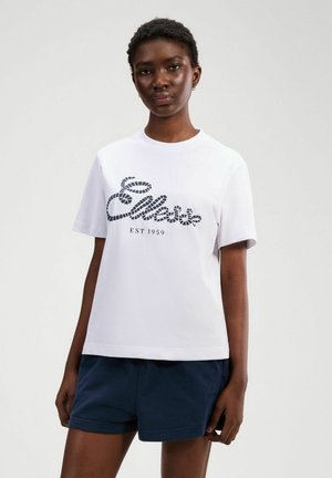 Młoda kobieta ubrana w białą koszulkę z krótkim rękawem z logo "Ellesse EST 1959" i granatowe szorty, stojąca na jednolitym tle.