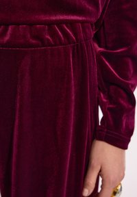 Zbliżenie na rękę osoby noszącej złotą i srebrną obrączkę, odpoczywającą obok burgundowego ubrania z weluru z elastycznym puntkiem.