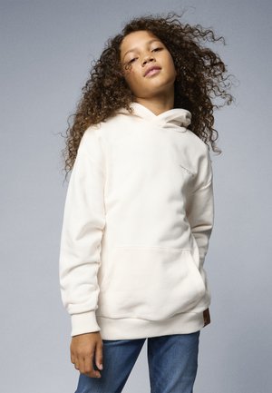 Hoodie couleur crème en tissu doux, avec une poche kangourou et une capuche à cordon, associé à un jeans bleu.