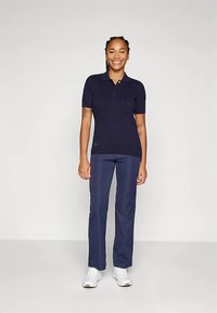 Χαμογελαστή γυναίκα με πλεγμένα μαλλιά, φορώντας ναυτικό μπλε πόλο με κοντά μανίκια, αντίστοιχες ναυτικές παντελόνες και λευκά αθλητικά παπούτσια, που στέκεται σε έναν απλό φόντο.