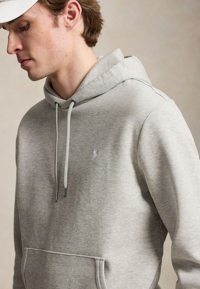Jeune homme portant un sweat à capuche gris clair avec cordons et poche kangourou, tourné vers la gauche sur un fond neutre.