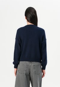 Oseba z dolgimi temnimi lasmi nosi mornarsko modro pulover in sivo jeans hlače, obrnjena proč proti enobarvni beli ozadju.