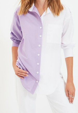 Femme portant une chemise à manches longues boutonnée, une moitié lavande et une moitié blanche, avec une poche blanche sur le côté droit.