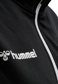  giacca nera con zip, caratterizzata da una superficie texturizzata, zip bianca e logo "hummel" in bianco sul davanti.