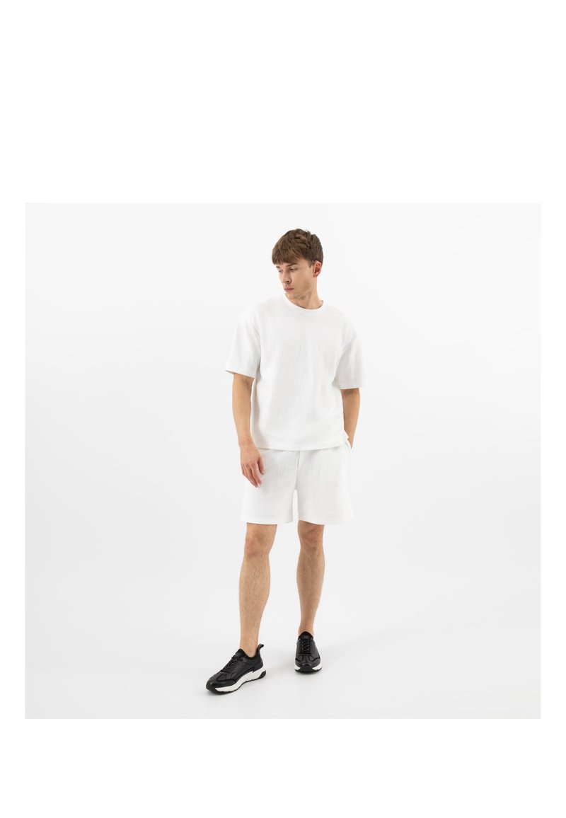 Mann steht und trägt ein weißes kurzärmeliges Hemd, weiße Shorts und schwarze Sportschuhe vor einem schlichten weißen Hintergrund.