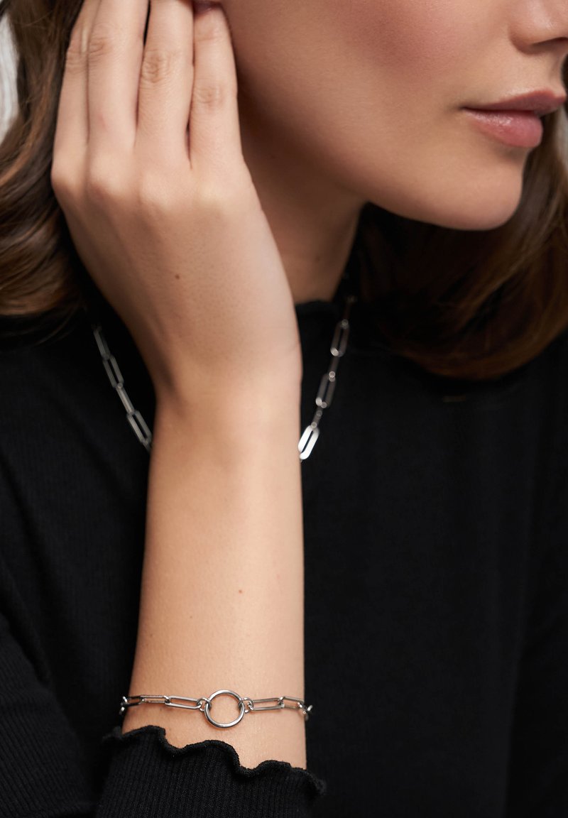 Sølv kædearmbånd med runde link accenter, kombineret med en lignende halskæde. Båret af en person mod en sort bluse, der fremhæver design detaljer.