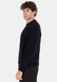 Maglione lavorato a maglia nero con scollo rotondo e polsini a coste. Tessuto morbido, vestibilità comoda e lunghezza moderata. Indossato da una modella in piedi di profilo.
